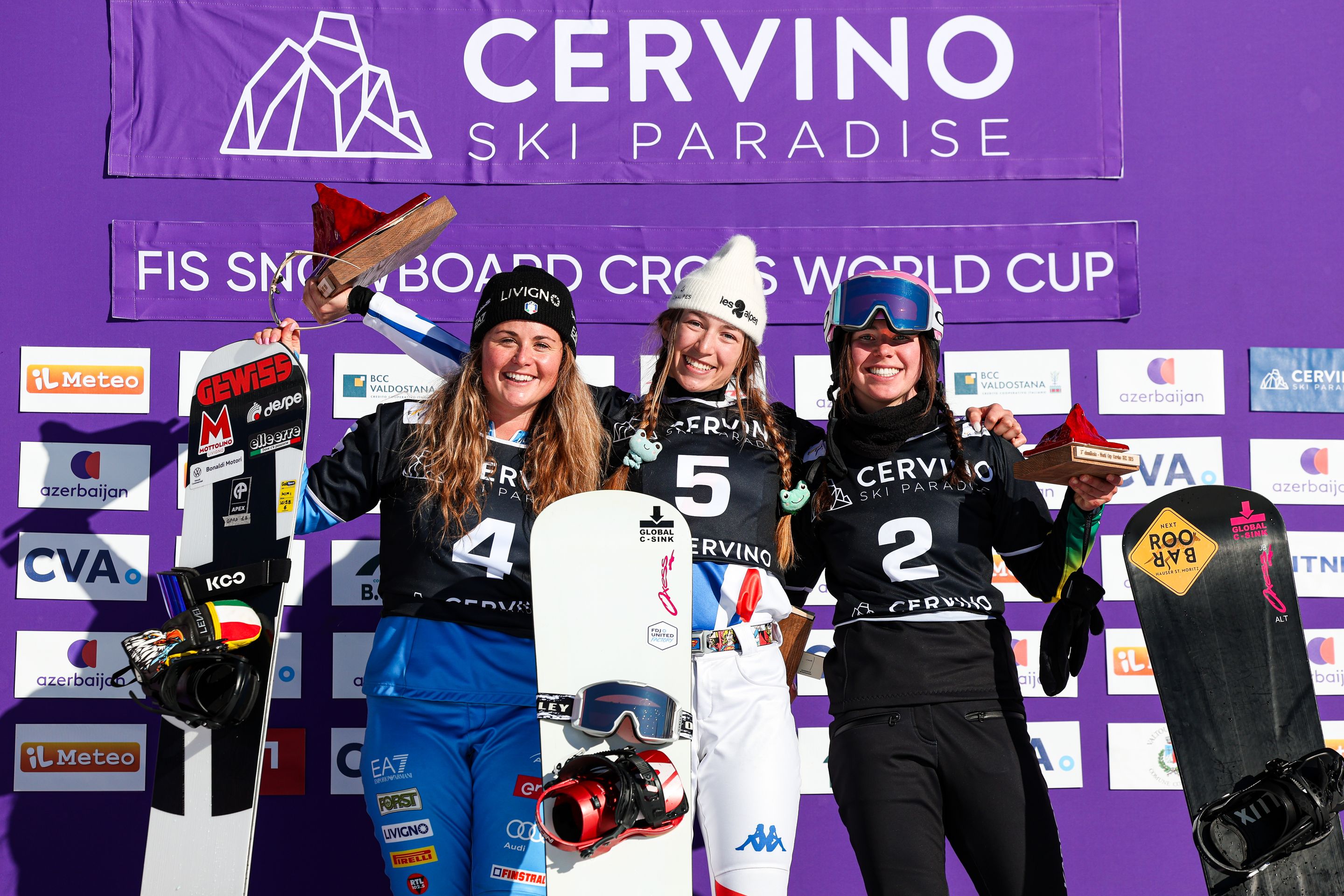 Three snowboarders in black bibs numbered 4, 5 and 2 celebrate in front of a purple backdrop reading “Cervino Ski Paradise” with sponsor logos