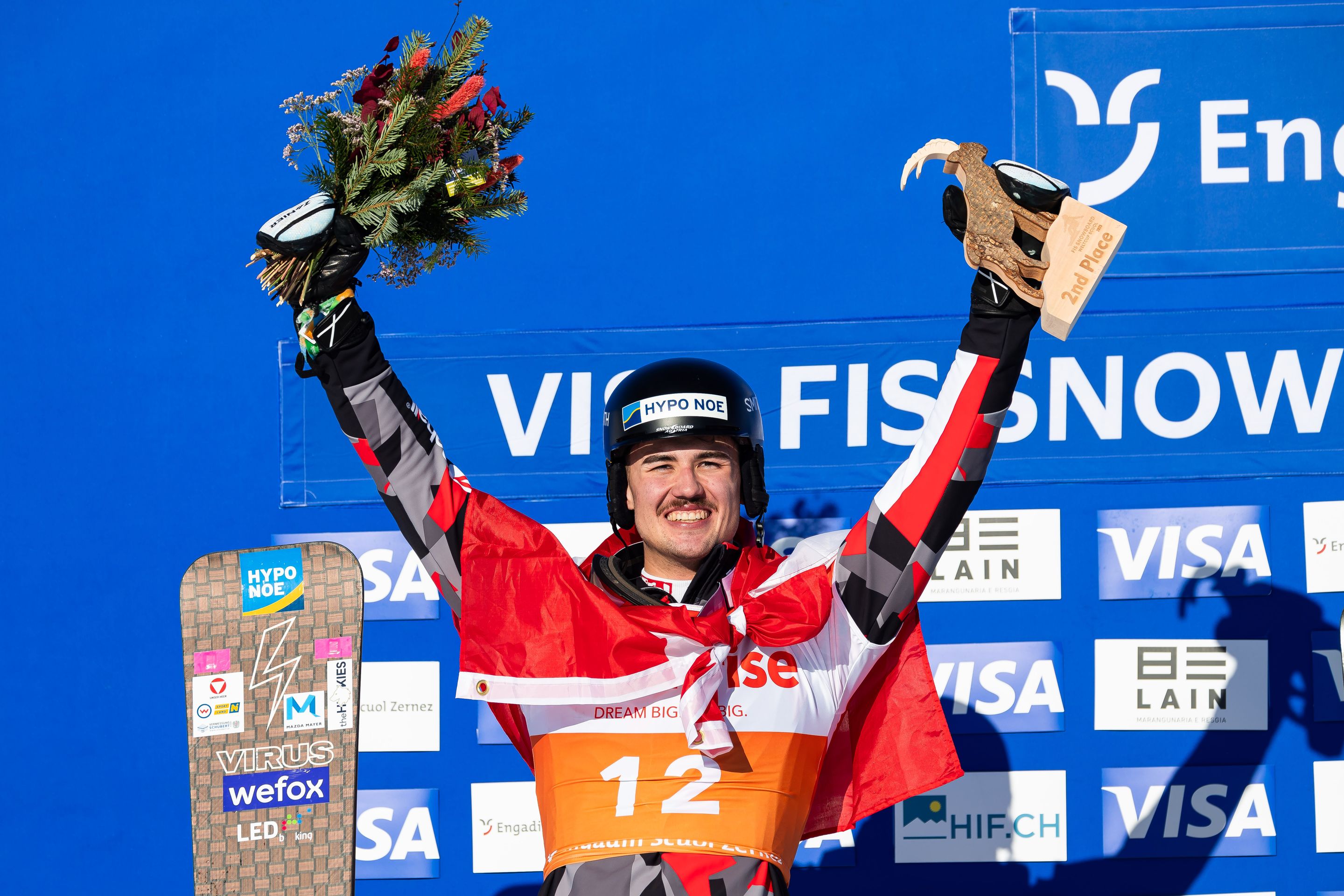 Snowboarder celebrates victory holding flowers and trophy, wearing a helmet and a red Swiss flag, standing on a podium.