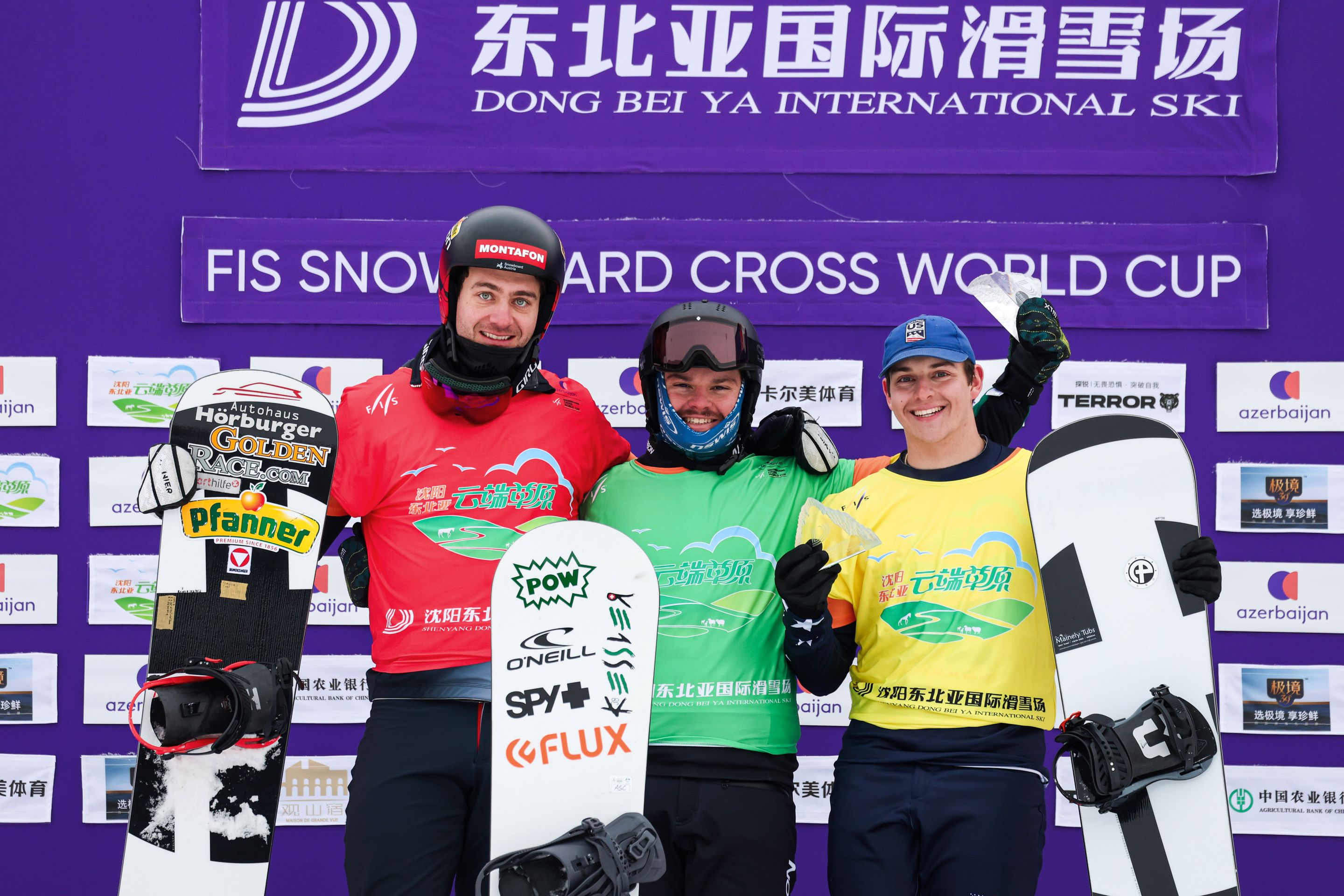 Three snowboarders in competition gear stand smiling with their snowboards in front of a backdrop at the FIS Snowboard Cross World Cup.