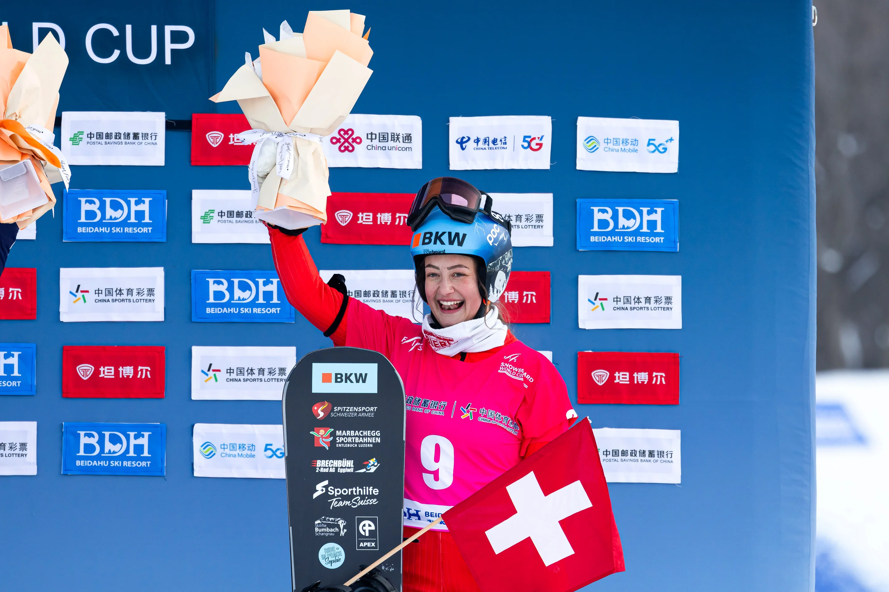 Sina Siegenthaler (SUI) celebrates on the podium in Beidahu, holding her snowboard and a Swiss flag and raising her gift in the air with a big smile