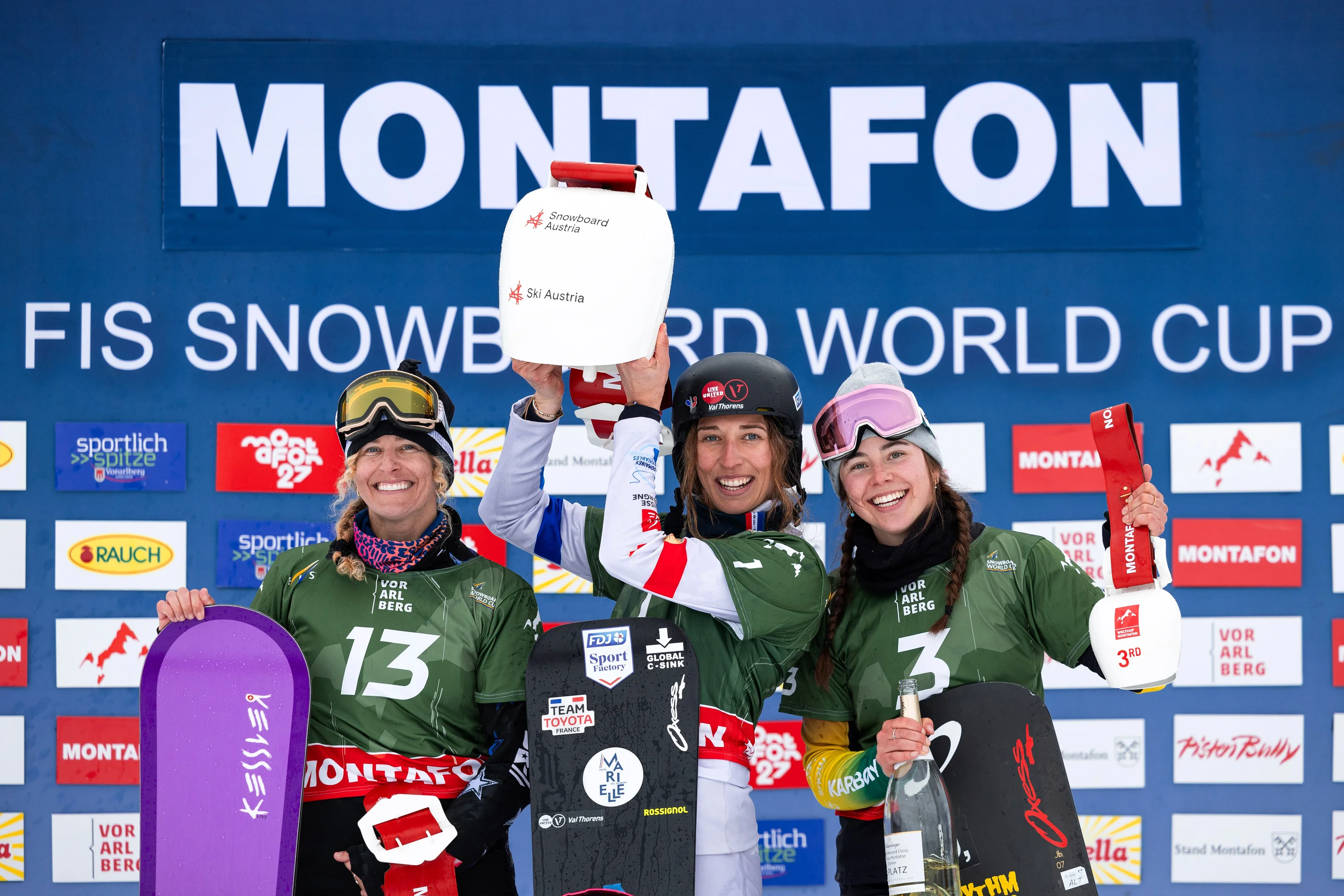 Three female snowboarders in green bibs stand together on the podium with their prizes, large white cowbells, in front of a blue backdrop reading ‘Montafon’