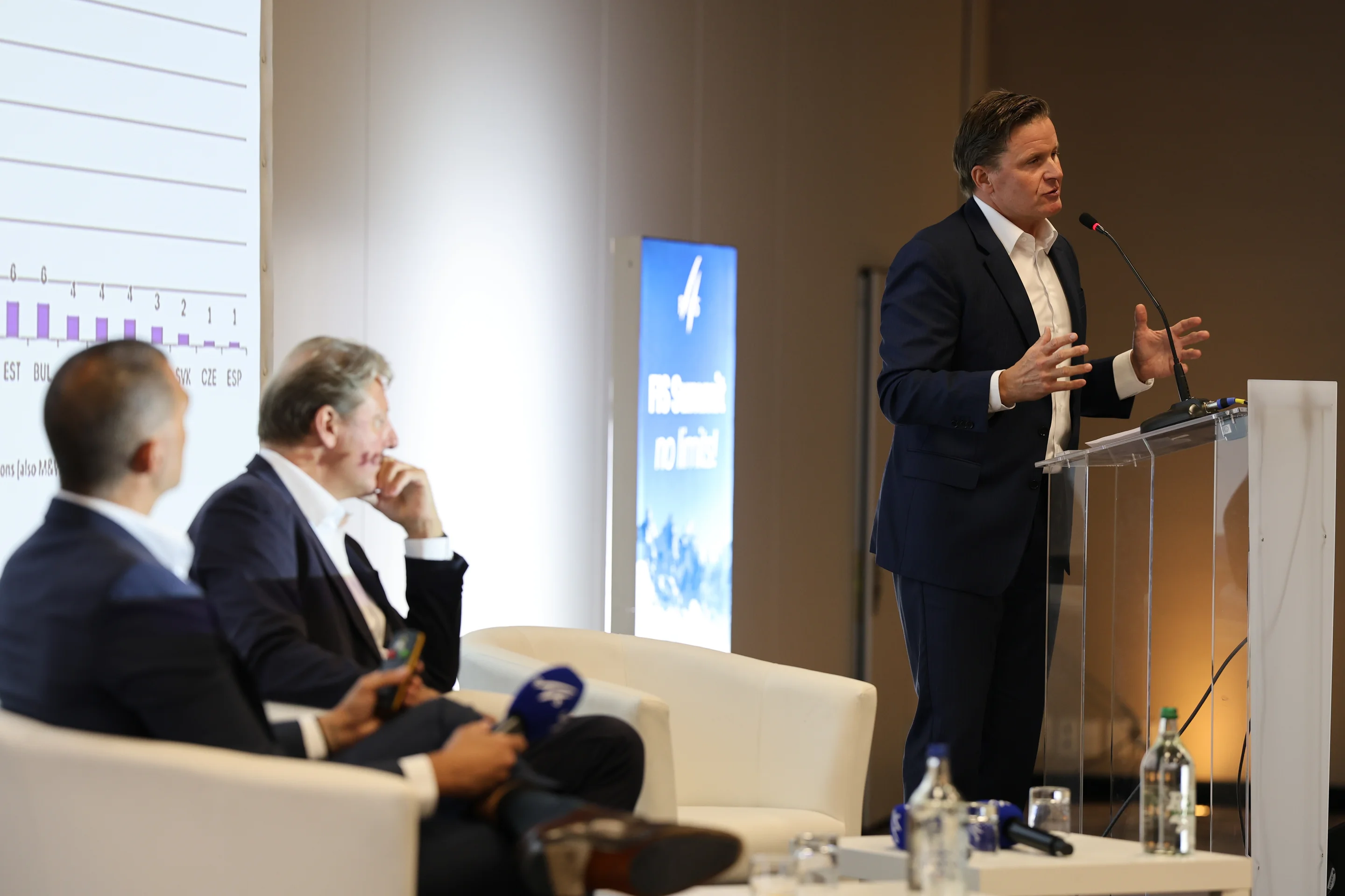 A man in a suit speaks at a podium during a conference, while two others sit and listen. A graph is visible on a screen beside them.