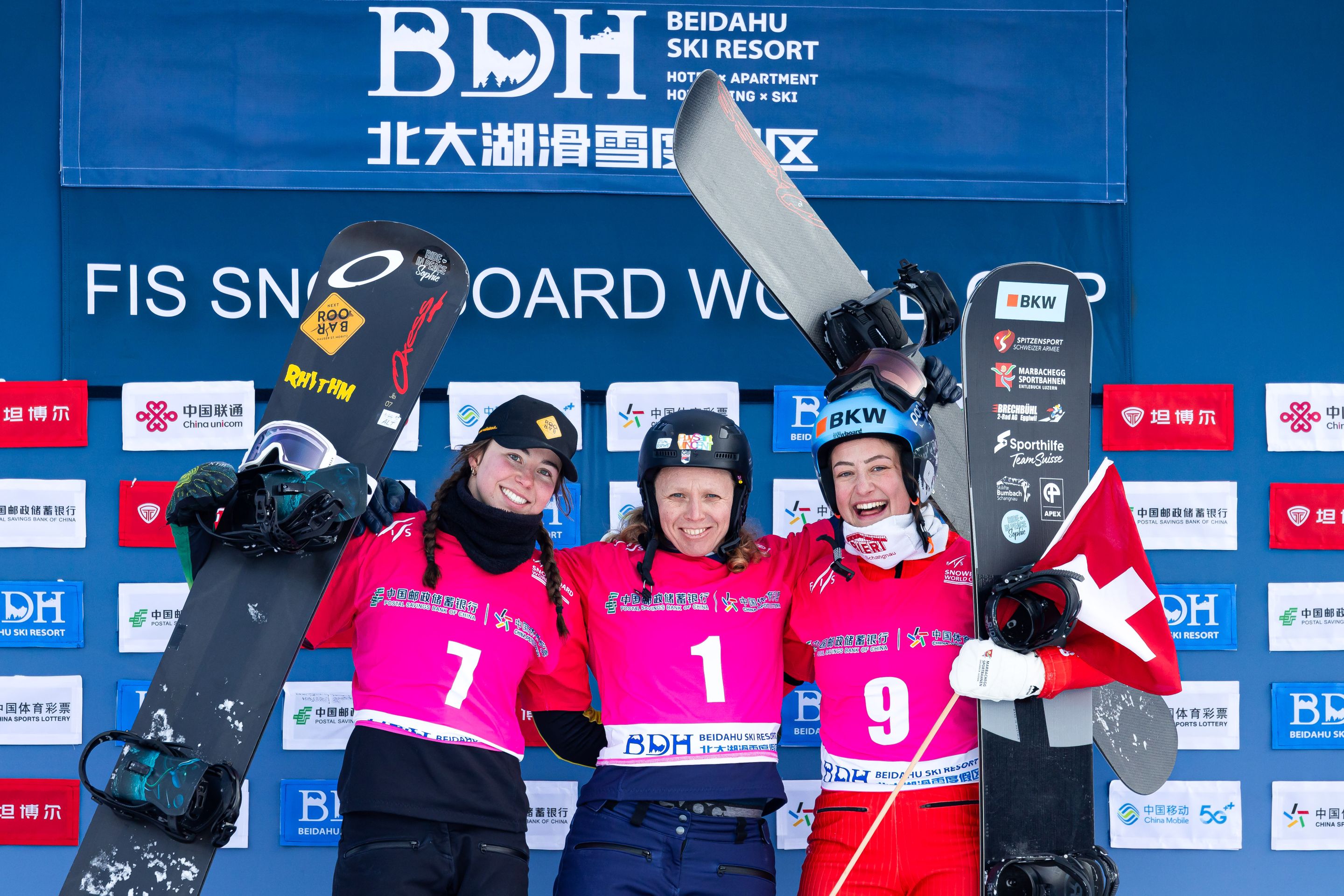 Three female snowboard athletes in pink bibs celebrate on the podium in Beidahu