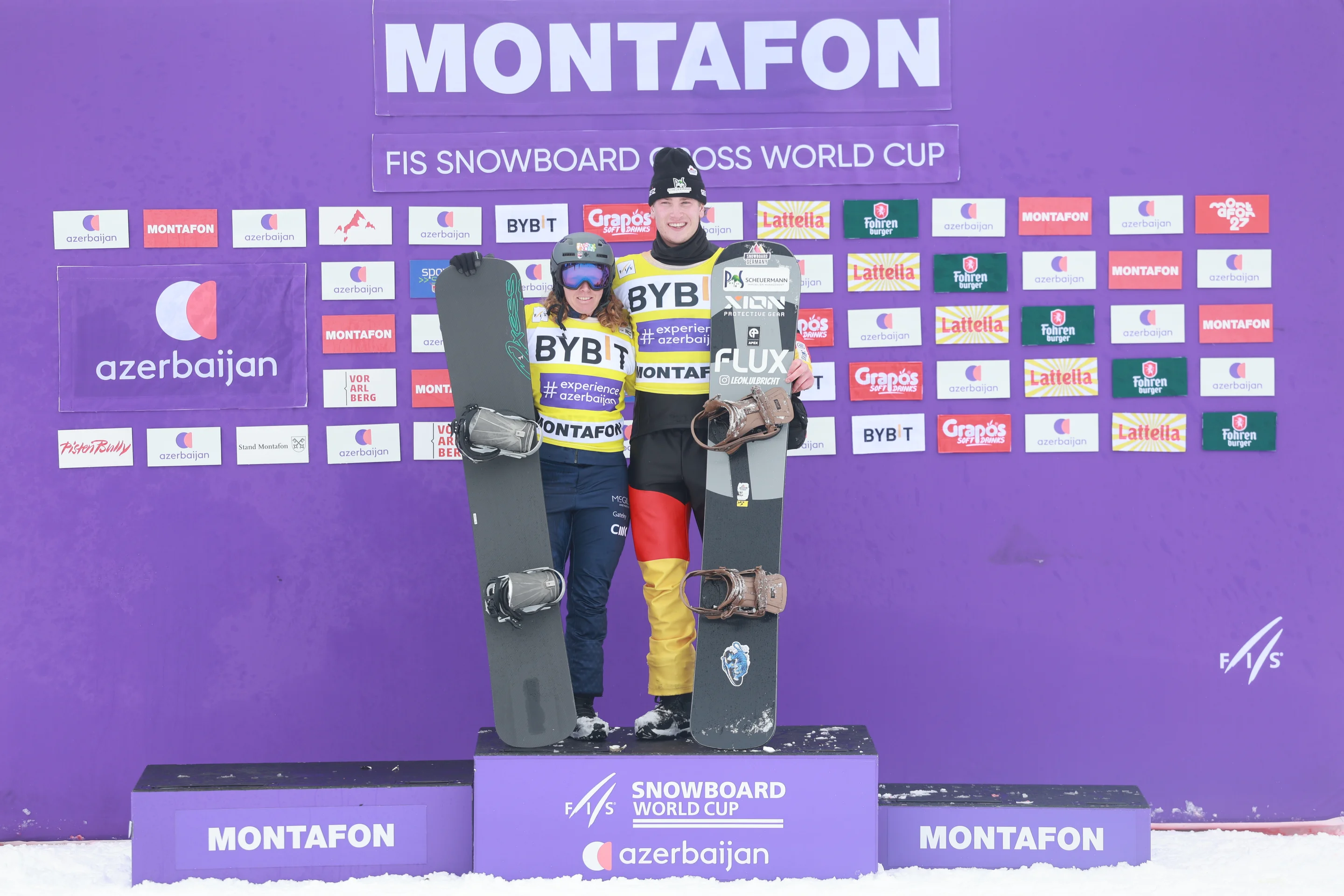 World Cup leaders Charlotte Bankes and Leon Ulbricht stand together on a purple podium in front of a purple ‘Monatfon’ backdrop. They wear yellow bibs and hold grey and black snowboards.