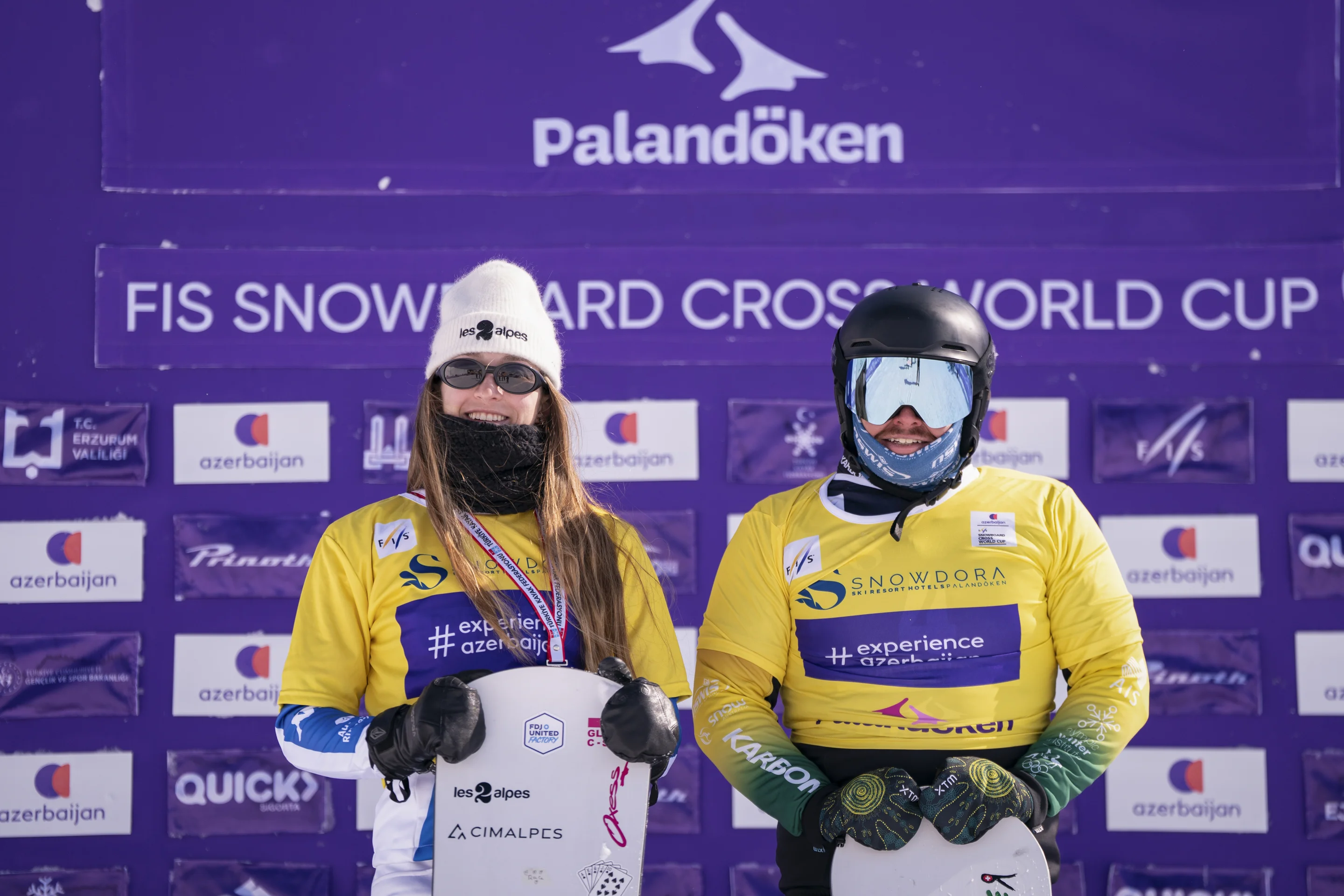 Lea Casta and Adam Lambert, in yellow bibs and smiling, stand in front of a purple backdrop as overall World Cup leaders
