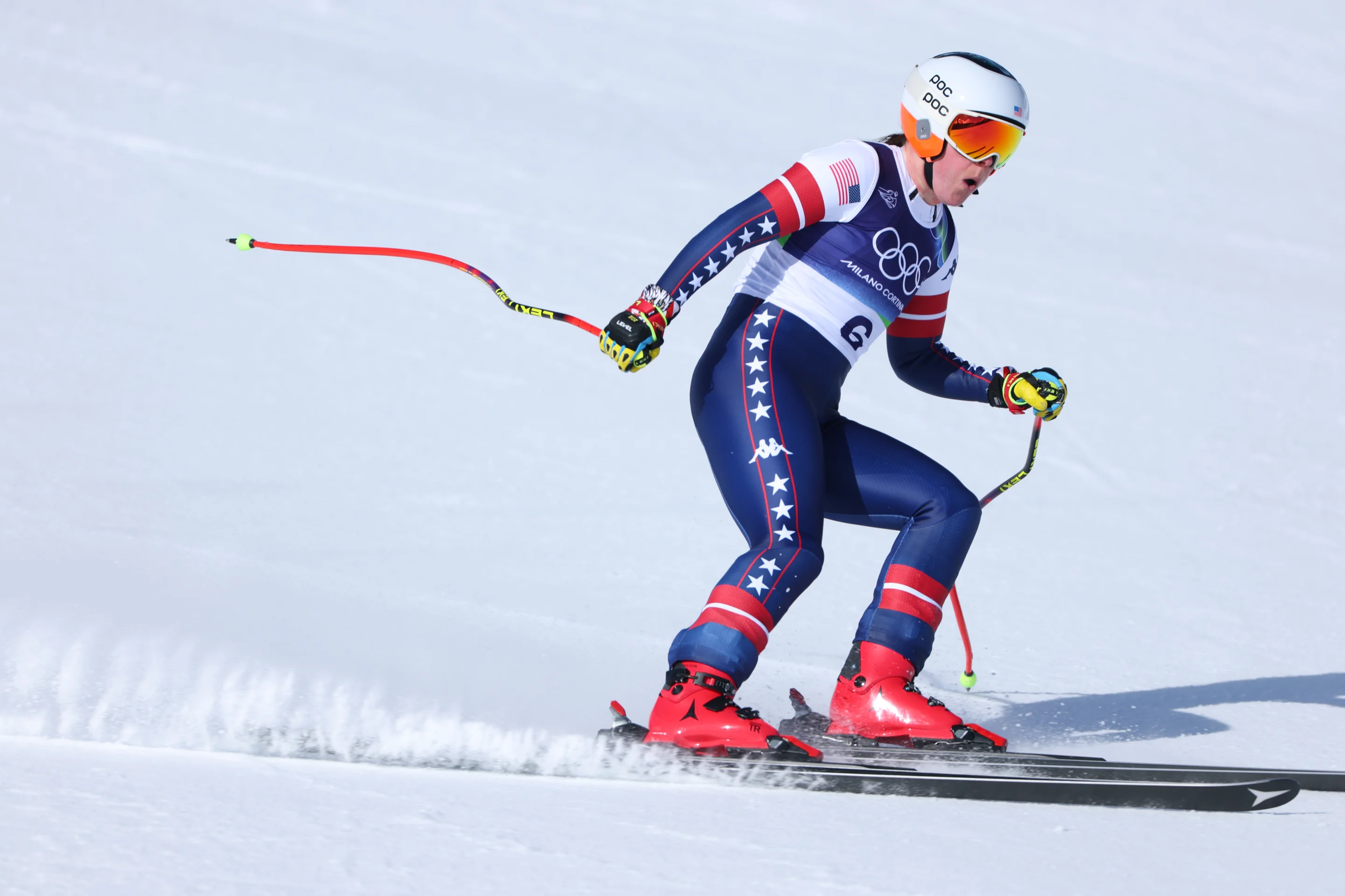 Ski racer in a star-patterned suit, skiing downhill on snow, wearing a helmet and reflective goggles, holding ski poles.