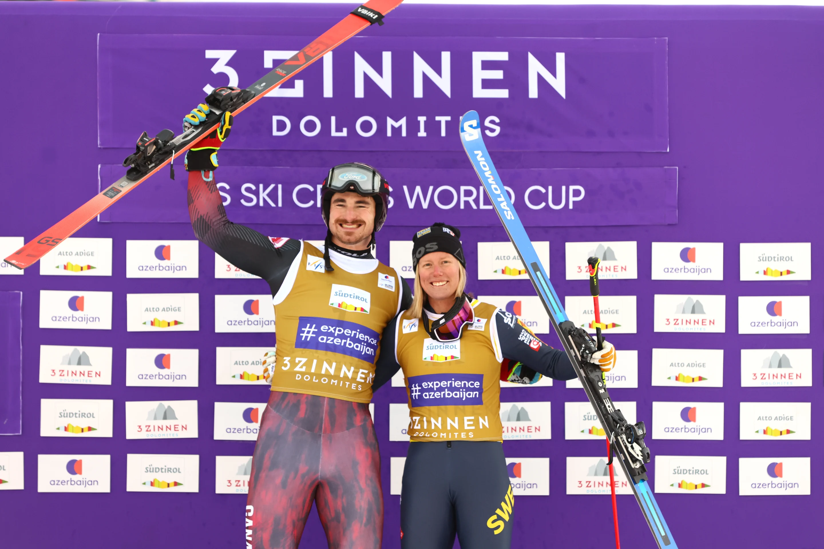 Wearing gold bibs over their ski suits, Reece Howden and Sandra Naeslund celebrate leading the World Cup standings by lifting their skis in the air, in front of a purple backdrop