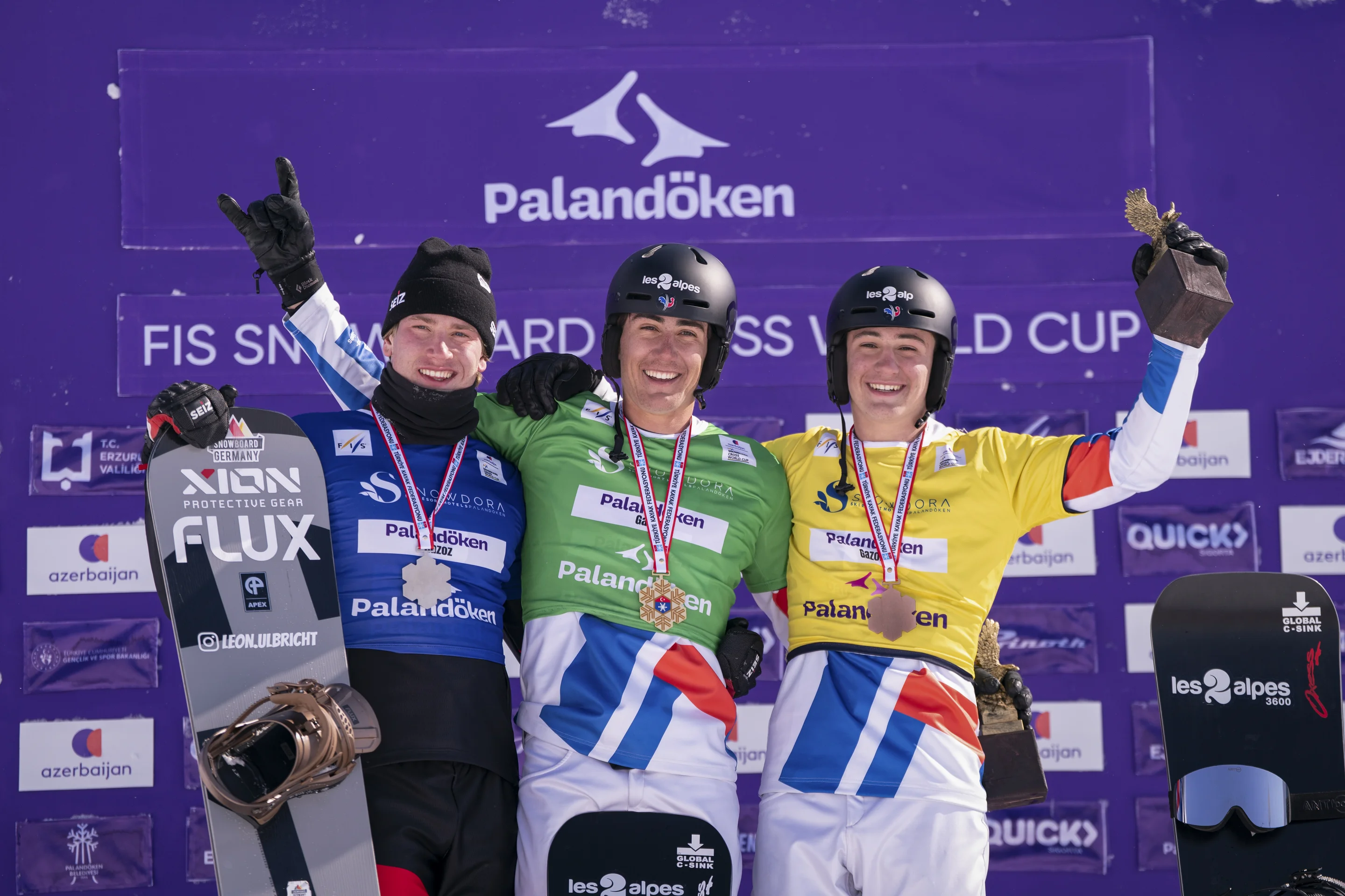 Wearing medals, Leon Ulbricht in a blue bib, Aidan Chollet in a green bib and Jonas Chollet in a yellow bib celebrate on a podium. Jonas Chollet is holding a trophy shaped like an eagle in his left hand up in the air.