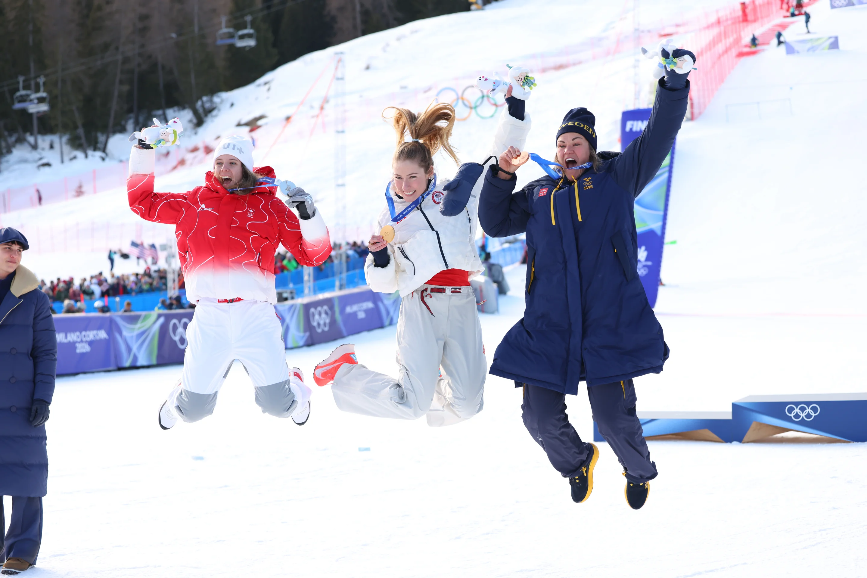 Three athletes in winter attire joyfully jump in the air on a snowy podium with Olympic rings in the background.