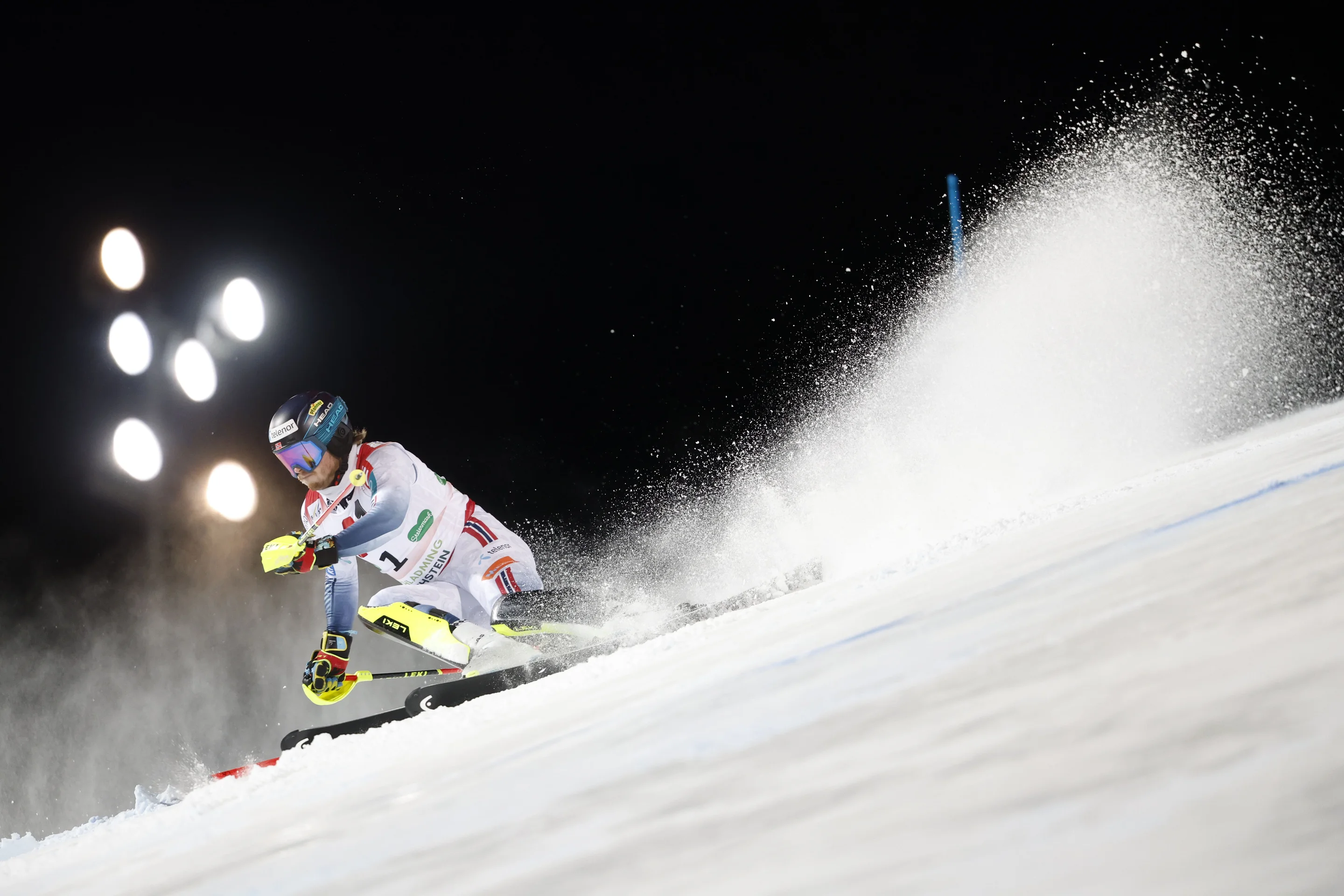 A skier in mid-turn on a snowy slope, illuminated by bright lights, with snow spraying in the air.