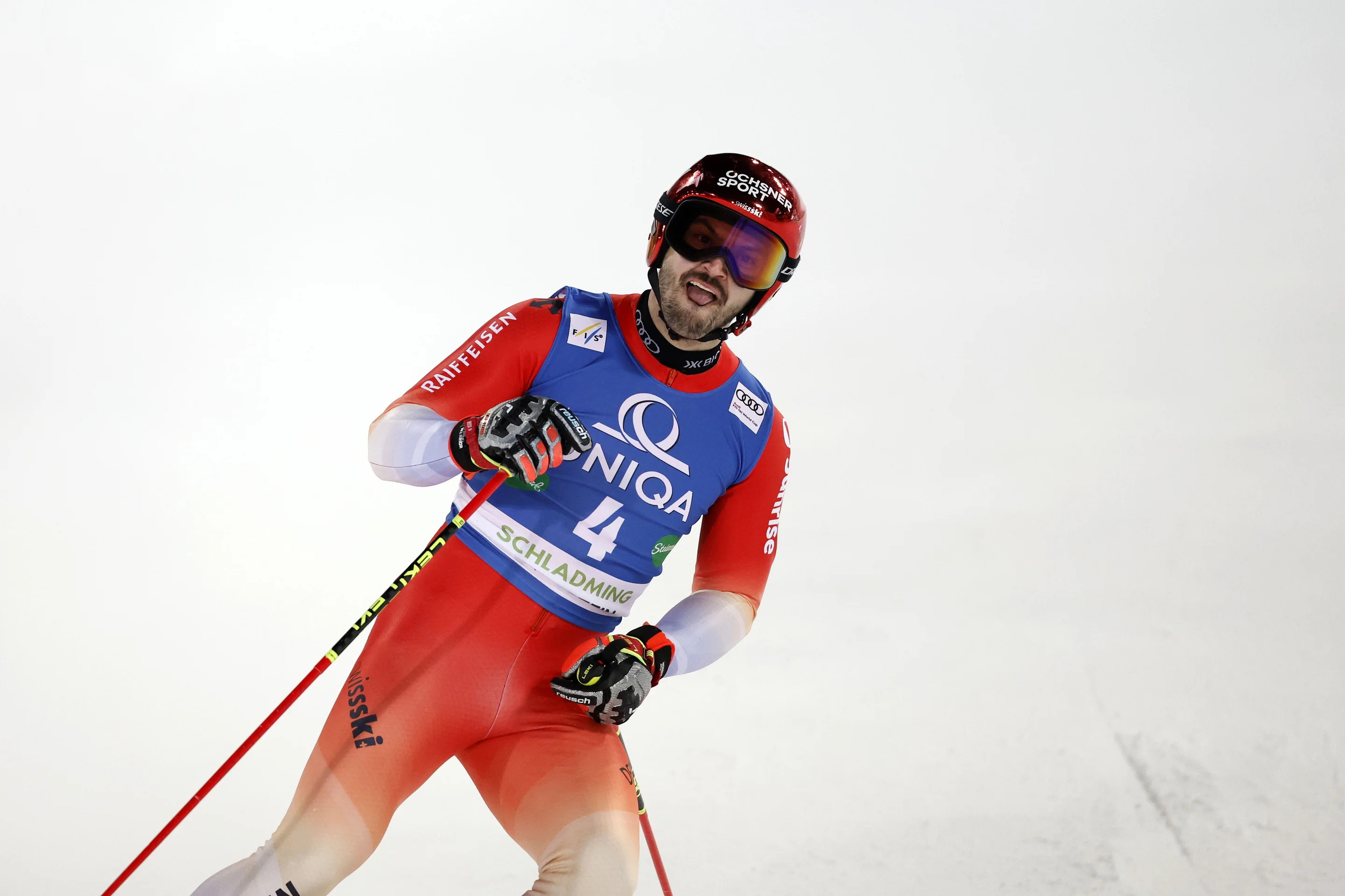 Ski racer in red and blue gear, wearing a helmet and goggles, holding ski poles, competing on a snowy slope.