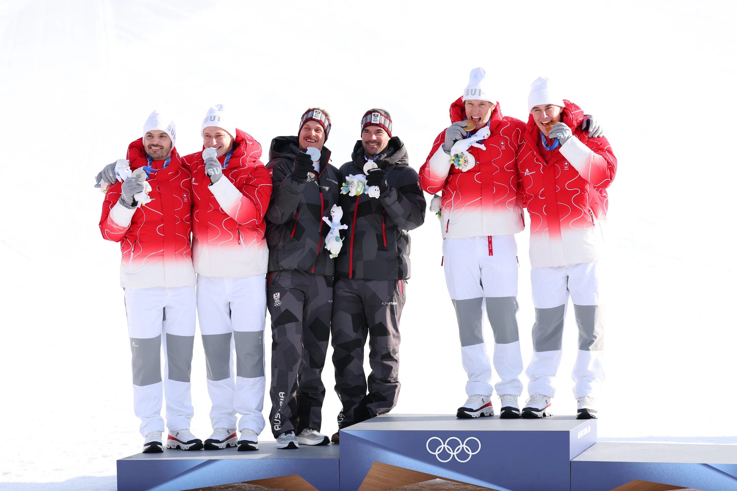 Six athletes in red and gray winter gear stand on a podium, each biting a medal, with snowy background and Olympic rings visible.