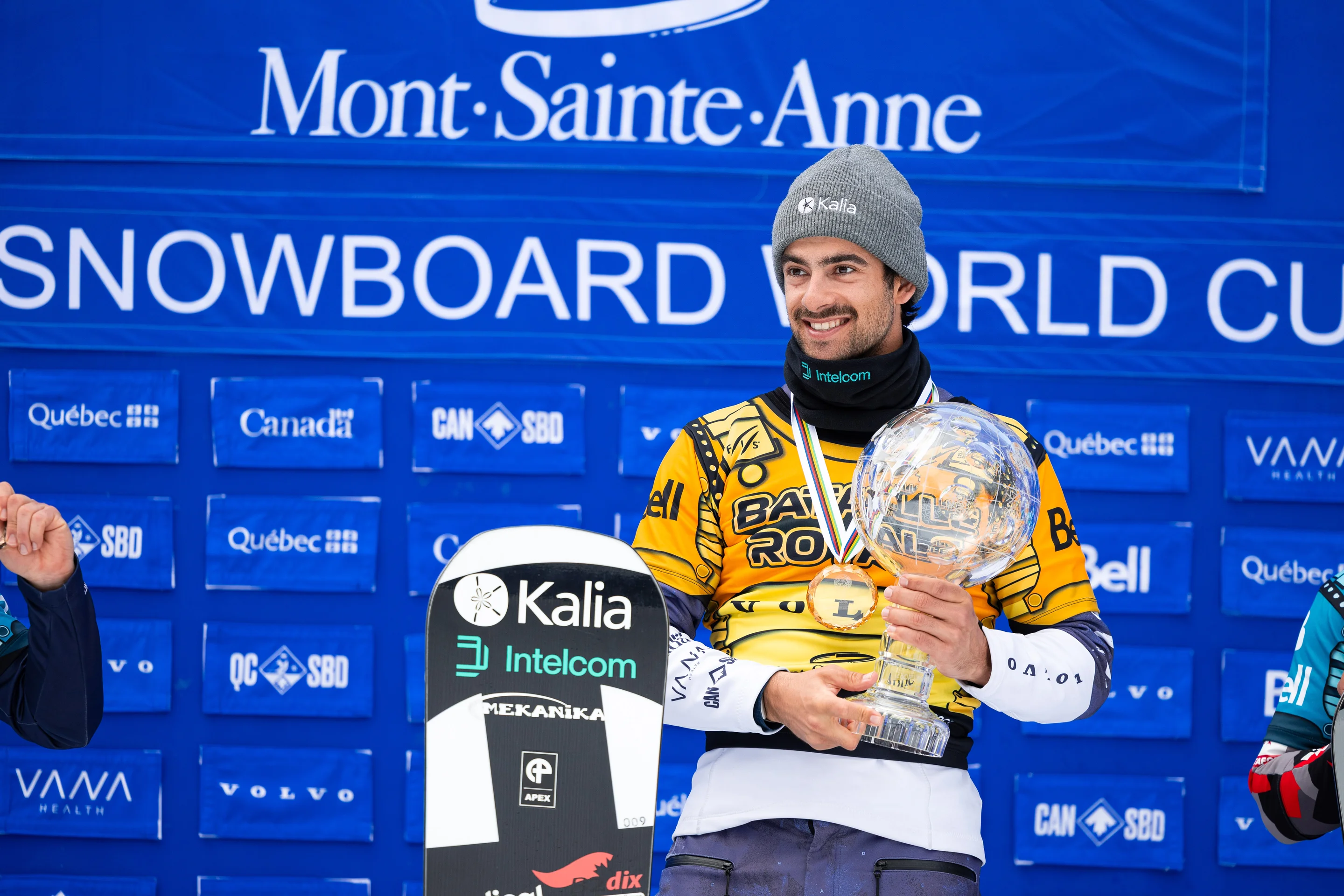 Wearing a yellow bib reading 'Bataille Royale' and a grey beanie hat, Eliot Grondin stands on a podium holding a trophy shaped like a crystal globe