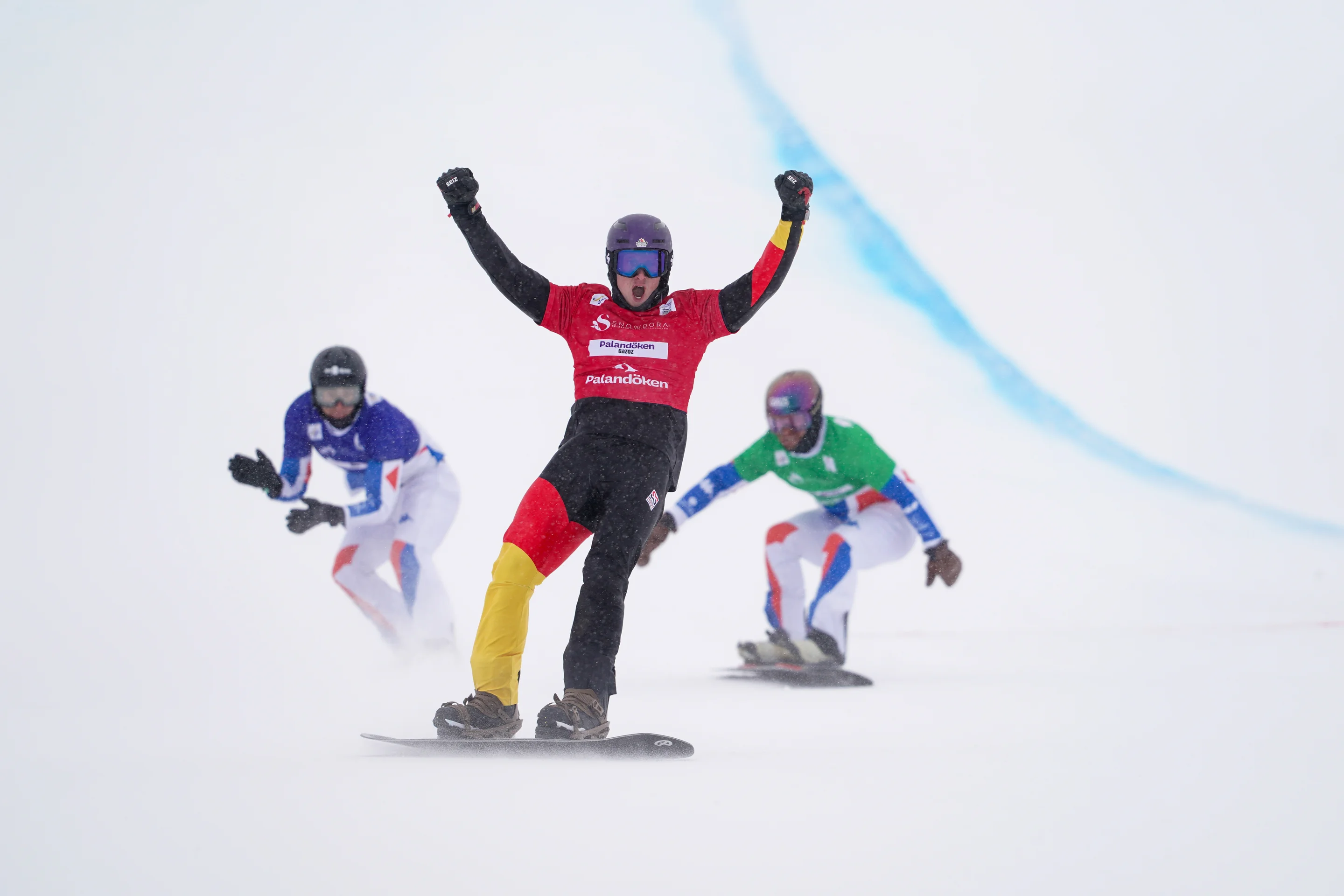 Snowboarder Leon Ulbricht raises his fists in the air in celebration after winning his race; two other snowboarders in French uniforms cross the line behind him