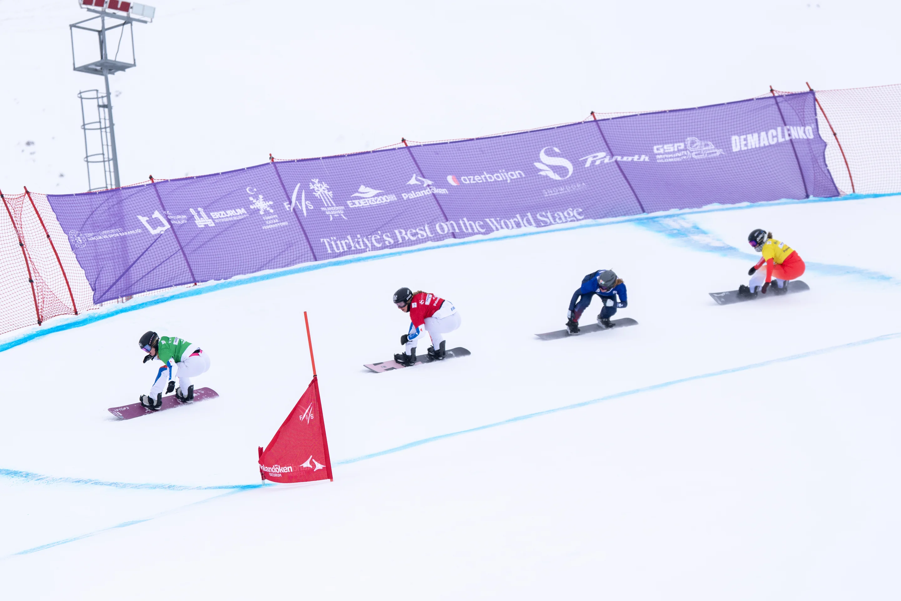 A line of racing snowboarders in coloured bibs are pictured racing around a turn