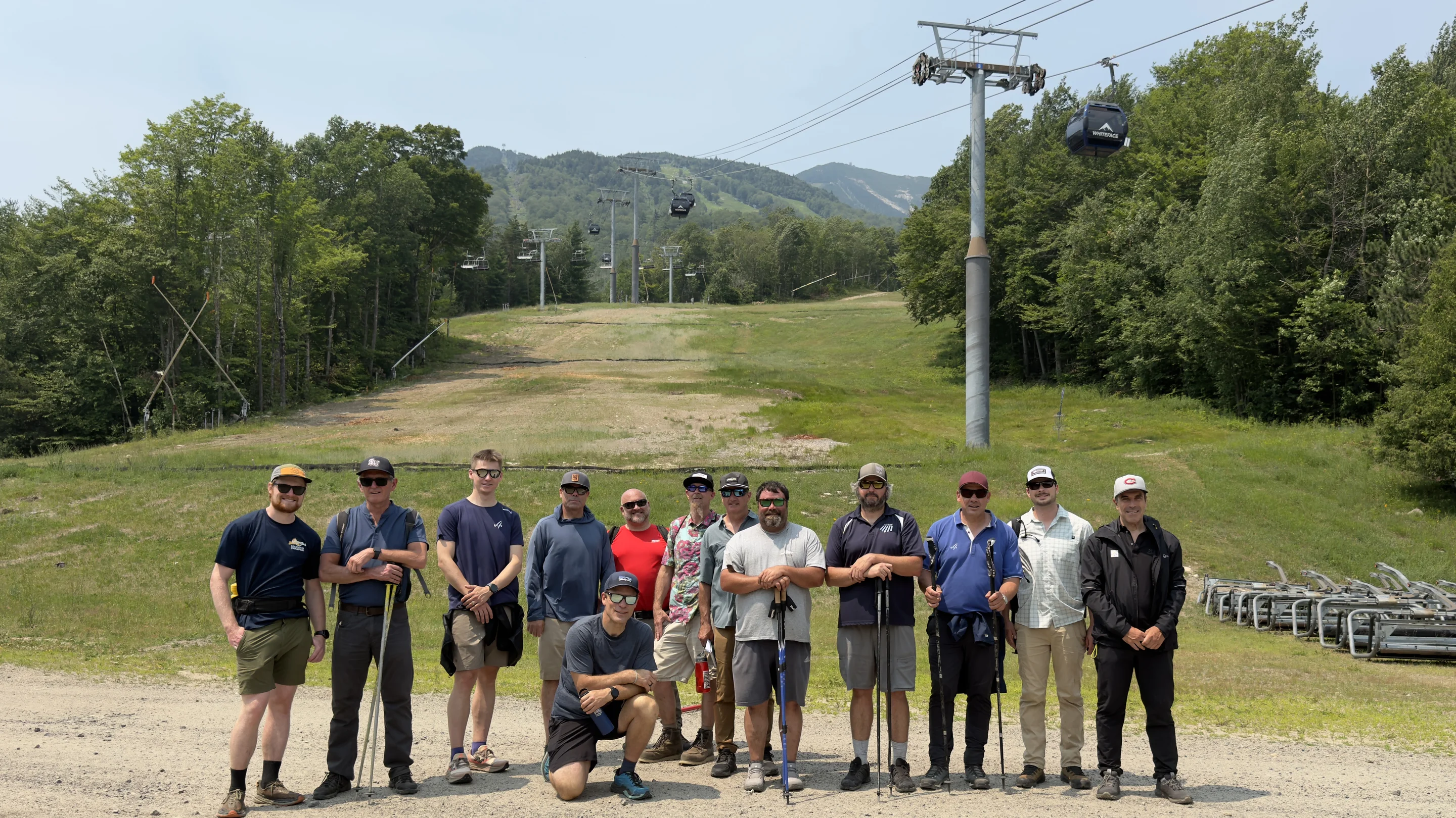 A group of 13 people with hiking gear stand in front of a ski lift on a green mountain slope with trees in the background.