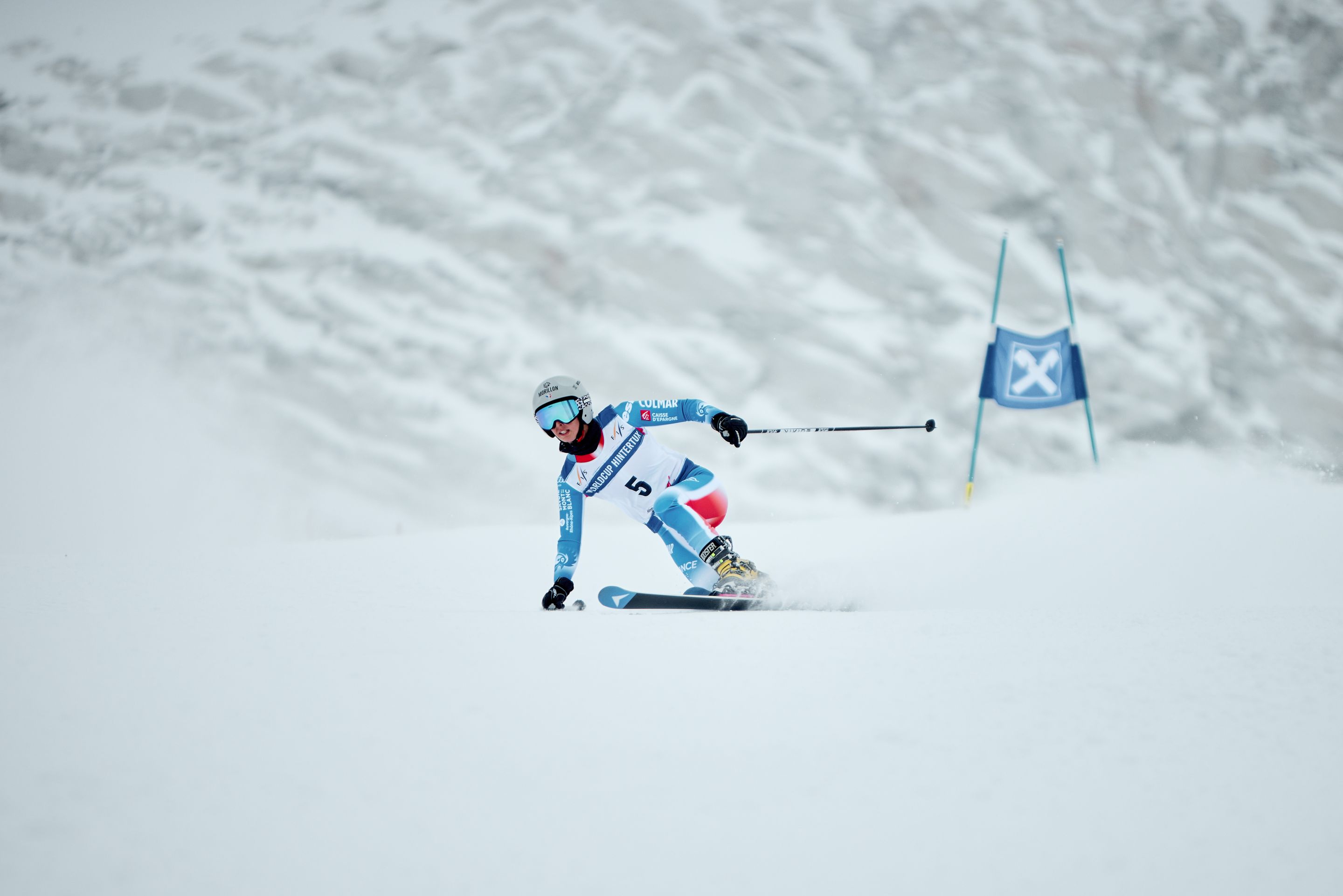 Skier in blue race suit (bib 5) carving a turn, kicking up snow near a blue slalom gate on a snowy mountain slope.