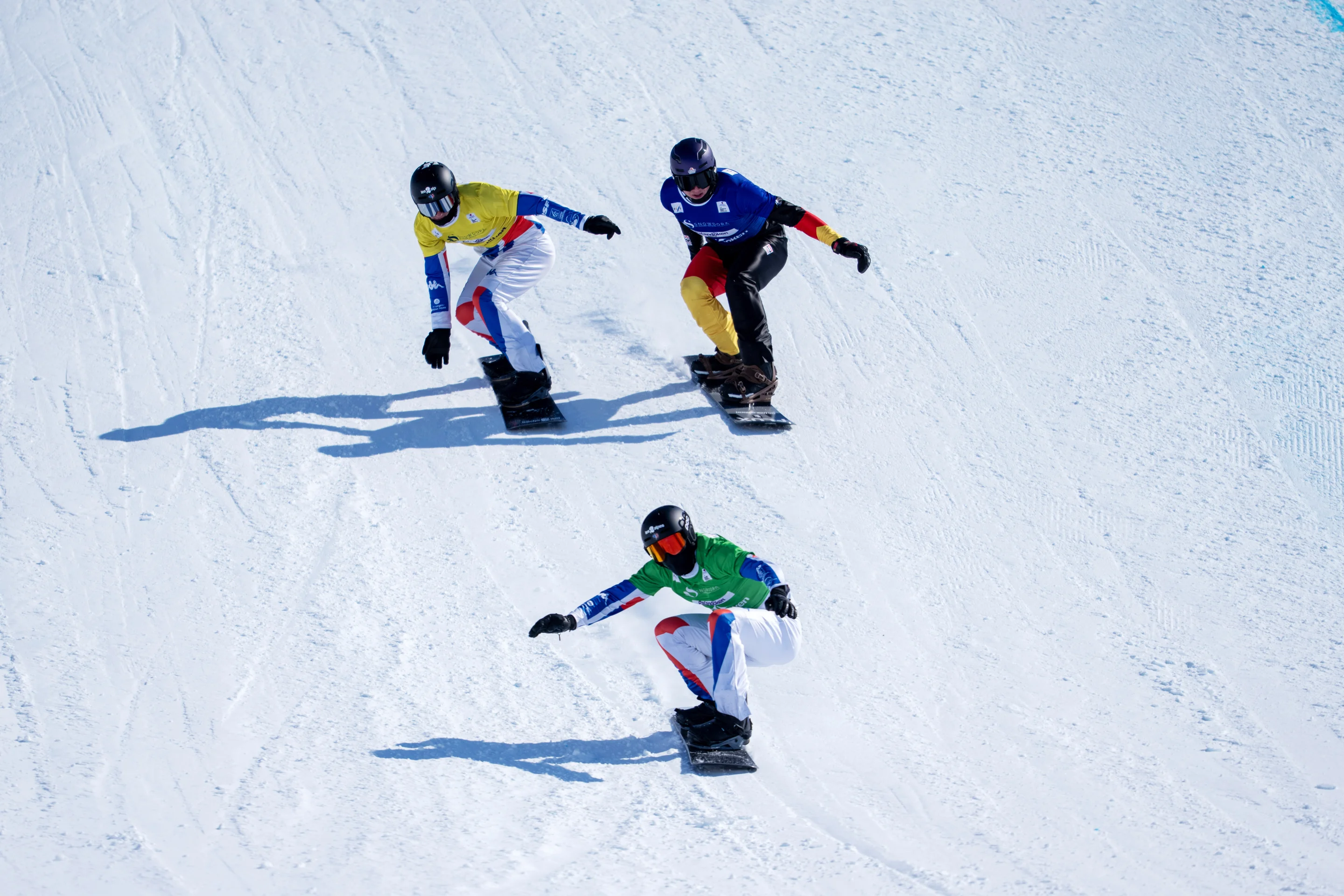 Aidan Chollet, in a green bib, leads Jonas Chollet in yellow and Leon Ulbricht in blue, all three snowboarders crouching for maximum speed on the snow