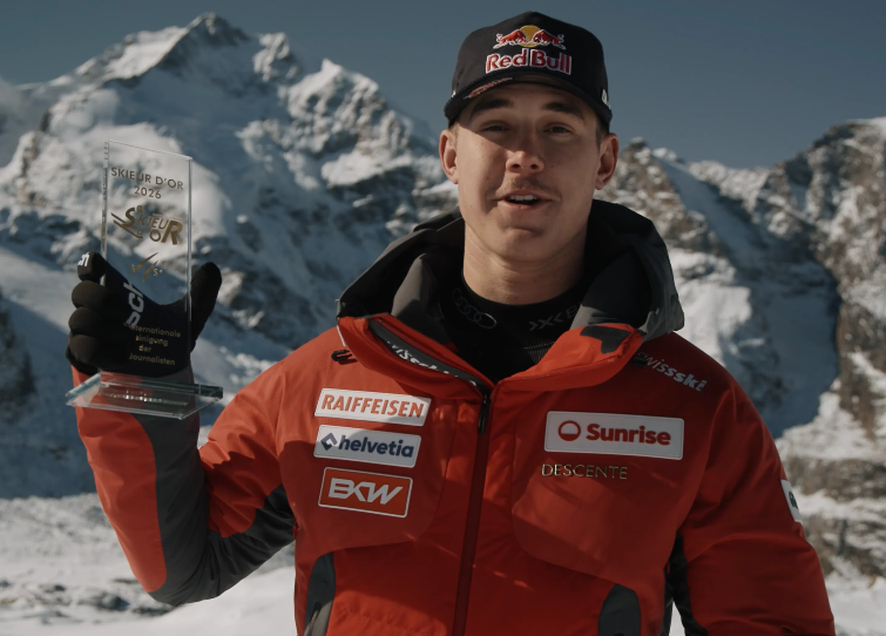 Franjo von Allmen, wearing a red Swiss team jacket and cap, holds up a glass trophy in front of snow-covered mountain peaks.