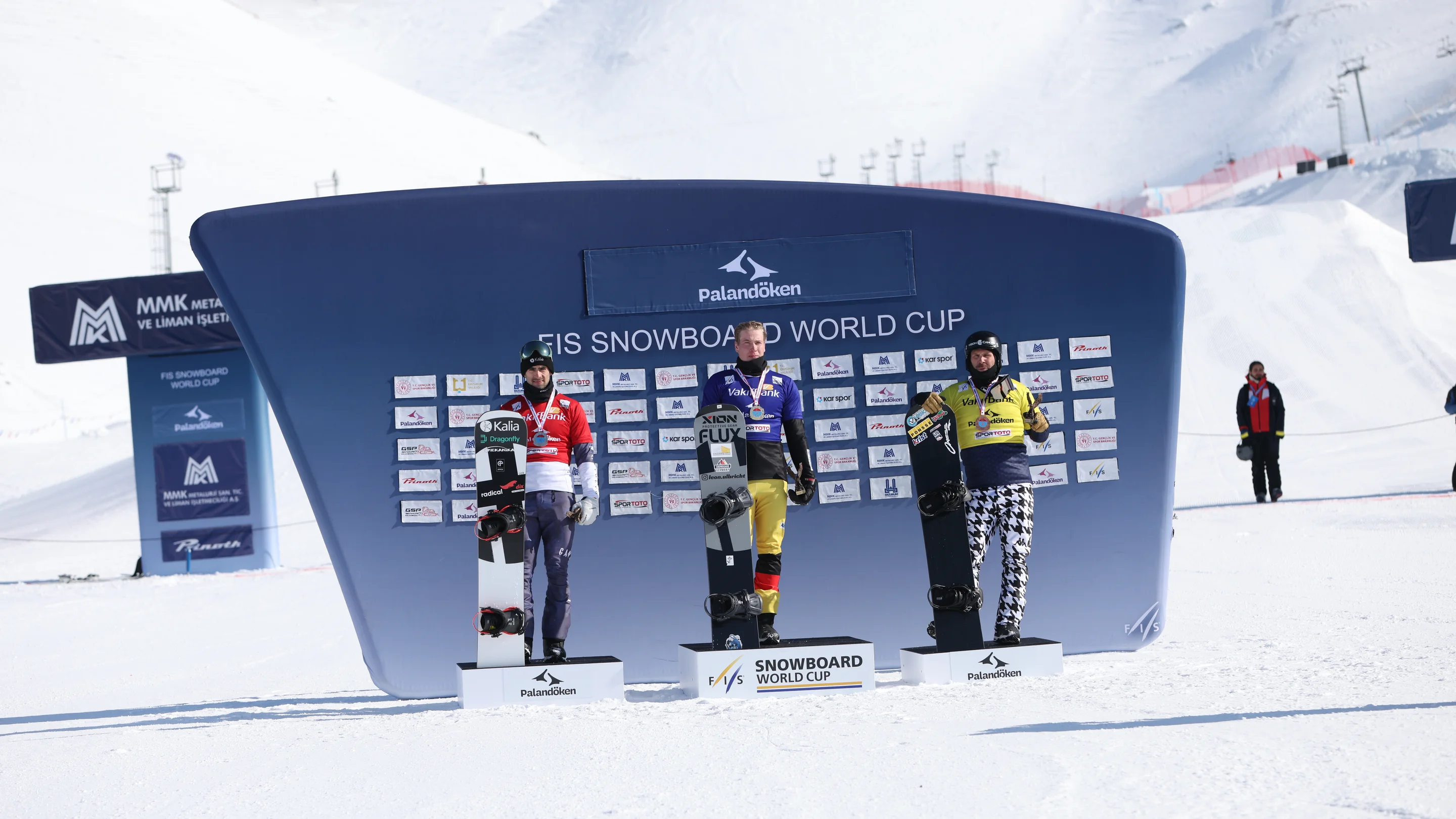 Three snowboarders stand on podiums in front of a blue backdrop and a snowy mountain