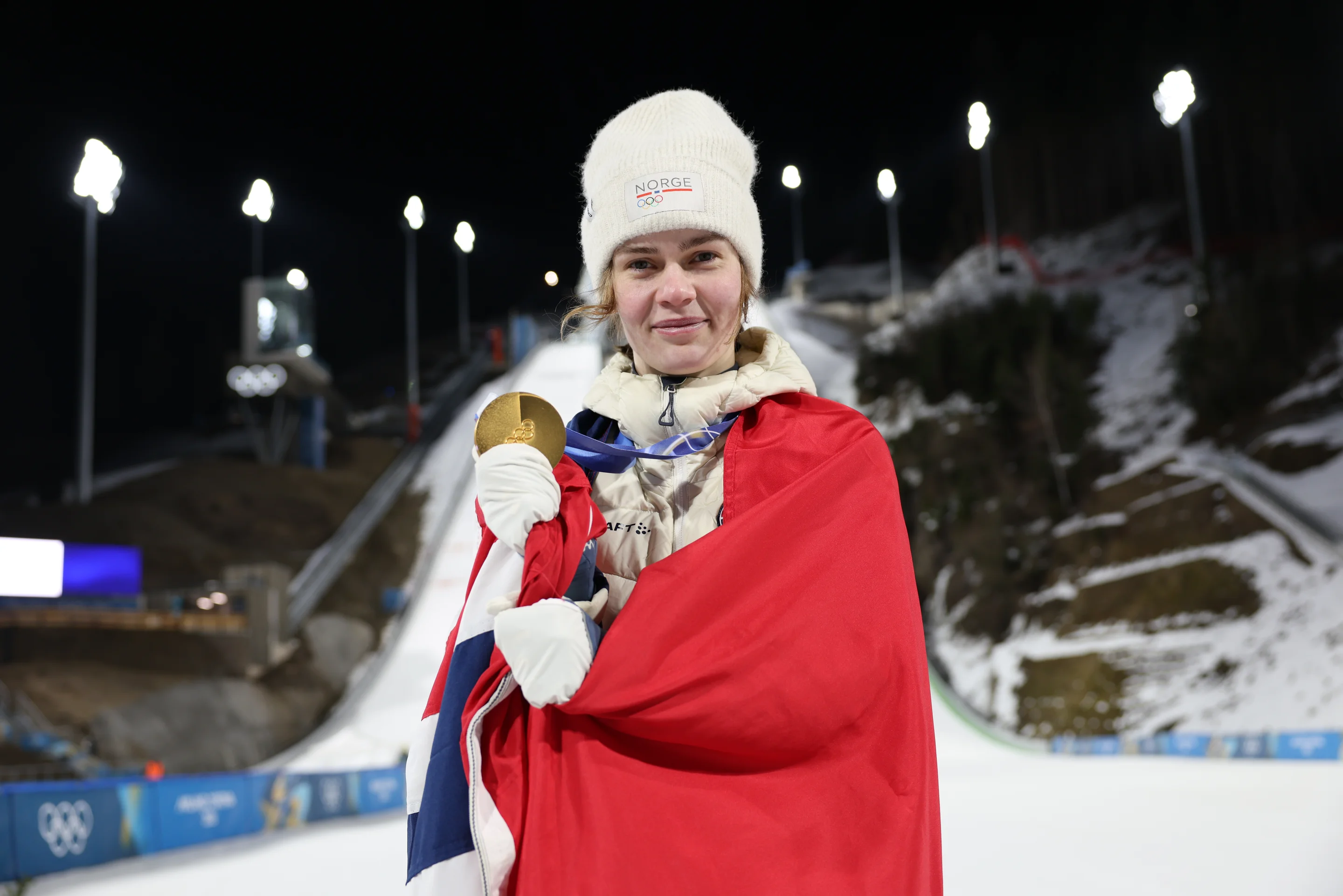 Athlete wrapped in a Norwegian flag holds a gold medal, standing on a snowy ski slope at night with bright lights in the background.