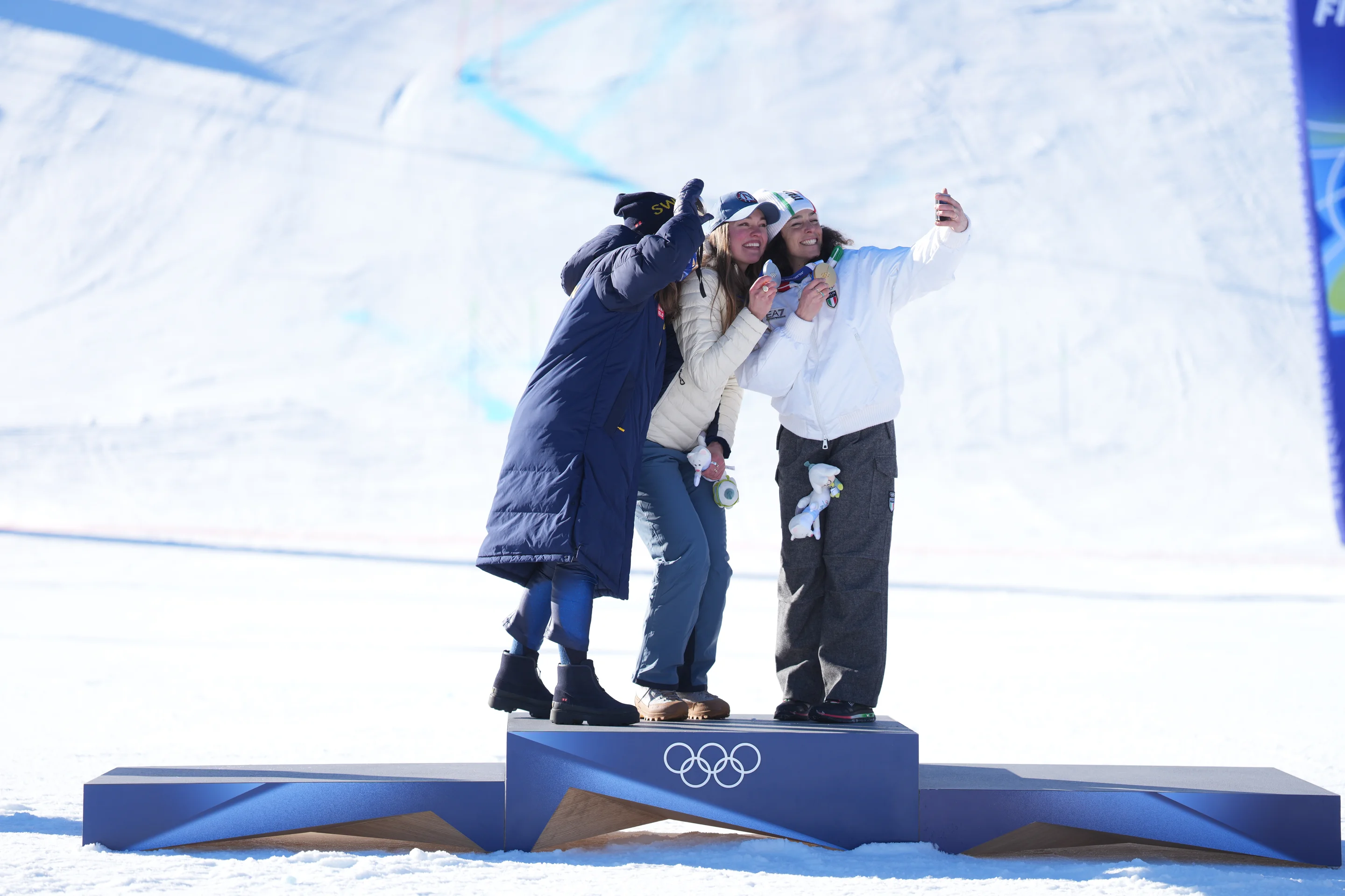 Three athletes on an Olympic podium taking a selfie together against a snowy background.