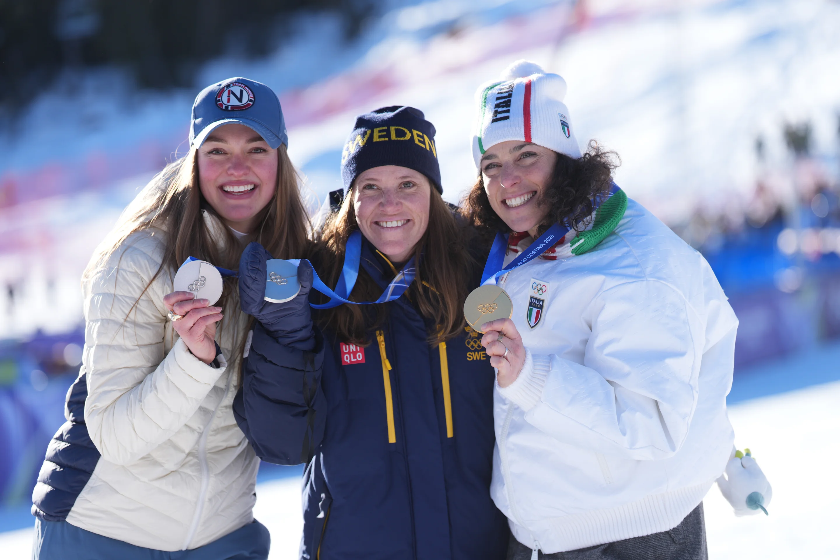 Three smiling athletes display their medals on a snowy winter day, wearing jackets and beanies.