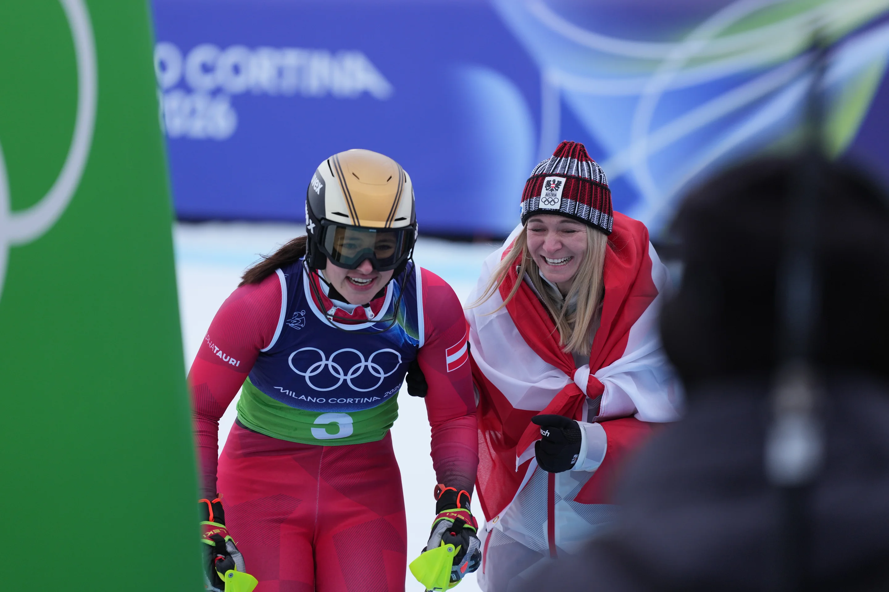 Two athletes in winter gear celebrating at a ski event; one wearing a helmet and goggles, the other draped in a flag and wearing a beanie.