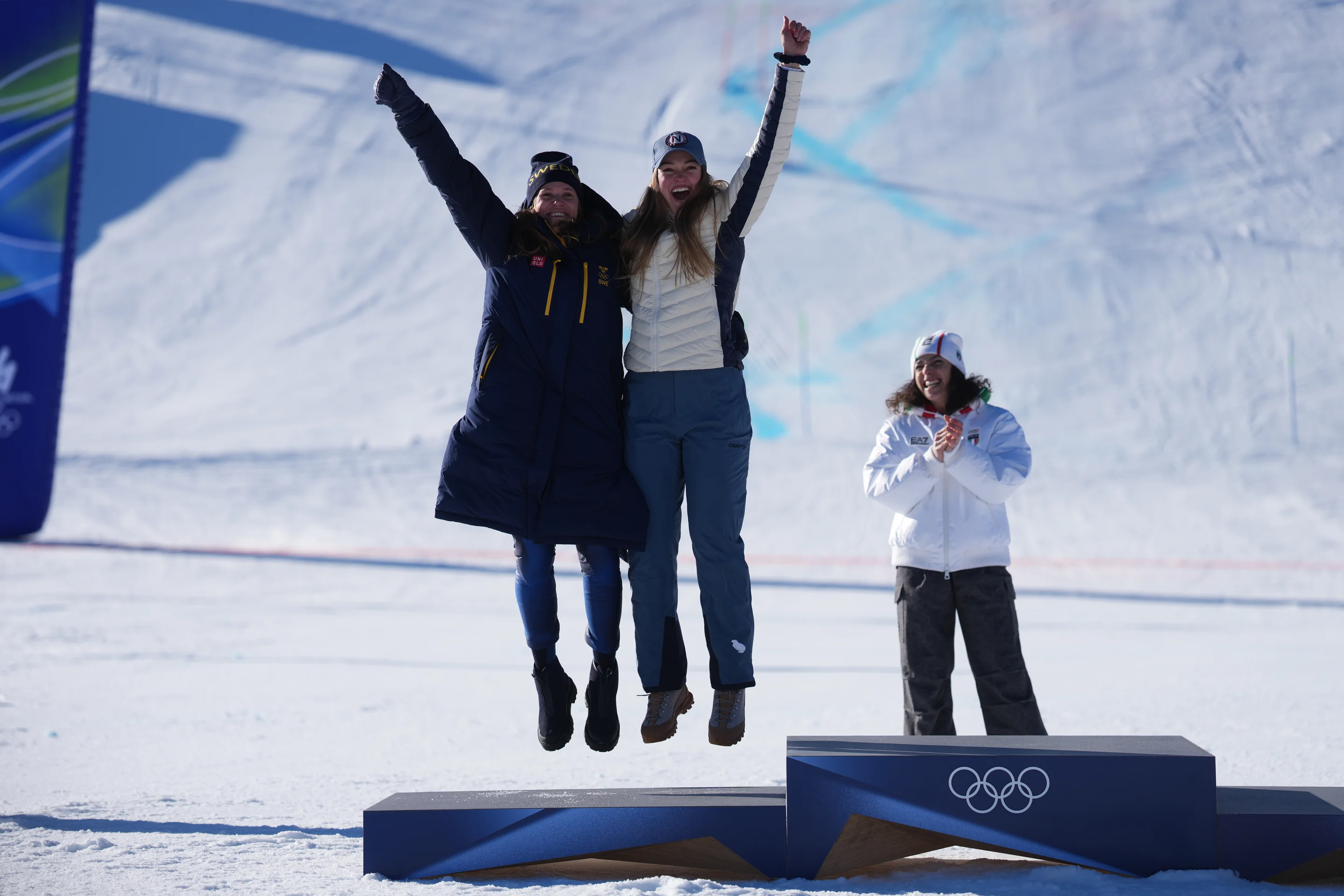 Two athletes joyfully jump on a podium in snowy conditions, celebrating, while another claps nearby, against a snowy backdrop.